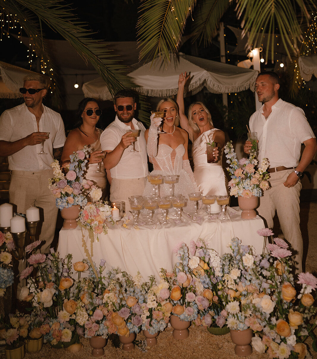 Group of people raising drinks from a table surrounded by flowers.